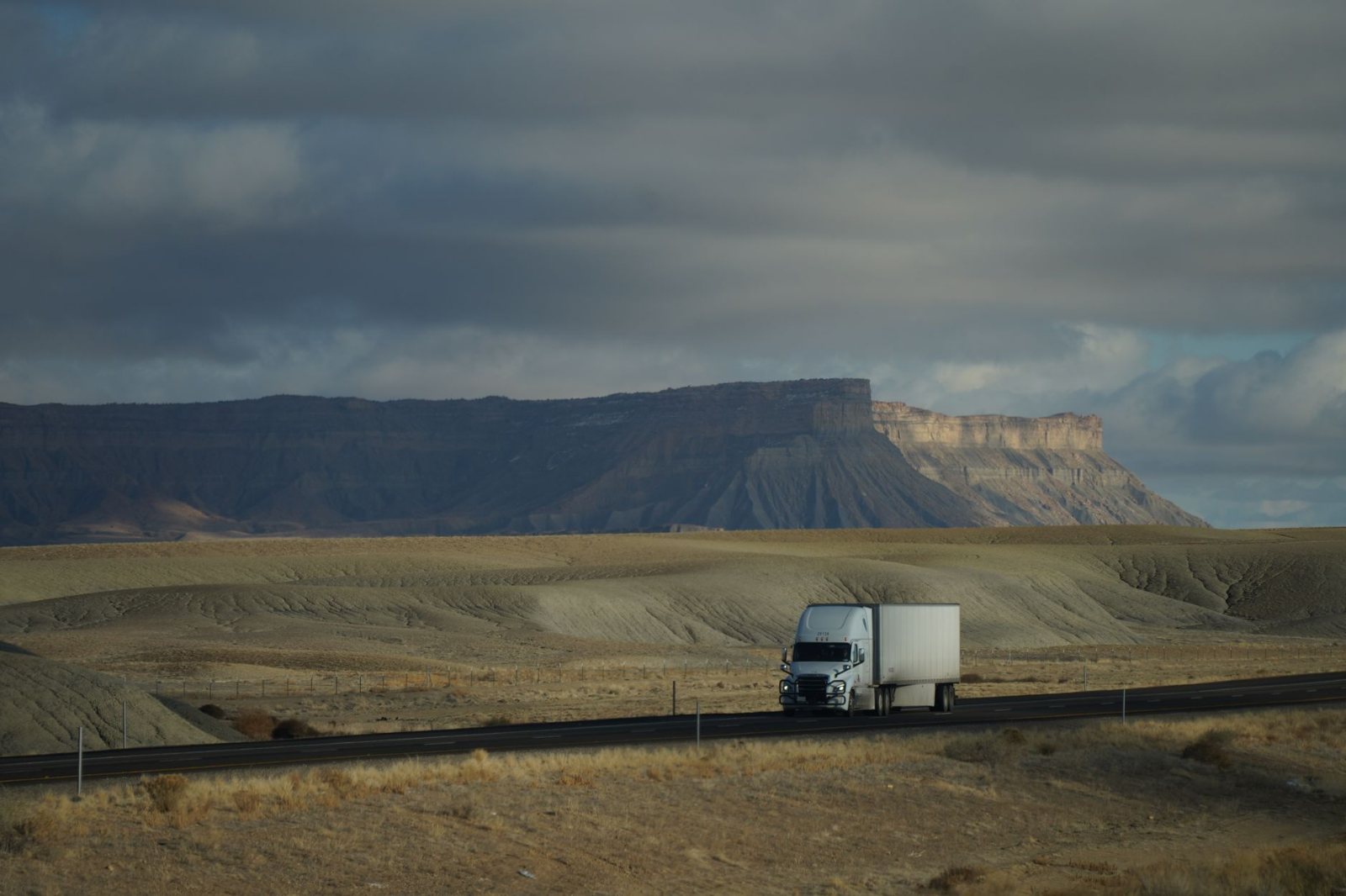 A tractor-trailer seen mid-run on an open interstate from a low three-quarter angle, lane markings streaking past, mirror and road ahead visible, flat grey overcast sky, no visible livery, no lens flare and no orange sunset grading